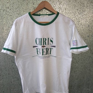 Puede incluir: Camiseta blanca con ribete verde y un gráfico de "Chris Evert" con el texto "This is a fine message touching body and refreshing mind. The exercise is in yourself."