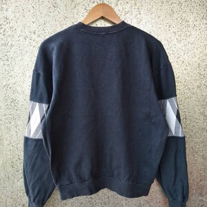 May include: A navy blue sweatshirt with a crew neck and long sleeves. The sleeves feature a white and black plaid pattern on the upper arm. The sweatshirt is hanging on a wooden hanger against a textured background.