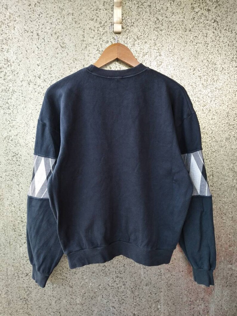 May include: A navy blue sweatshirt with a crew neck and long sleeves. The sleeves feature a white and black plaid pattern on the upper arm. The sweatshirt is hanging on a wooden hanger against a textured background.