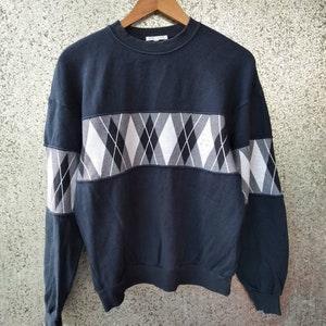 May include: A navy blue sweatshirt with a white and black argyle pattern across the chest and upper sleeves. The sweatshirt is hanging on a wooden hanger against a textured background.