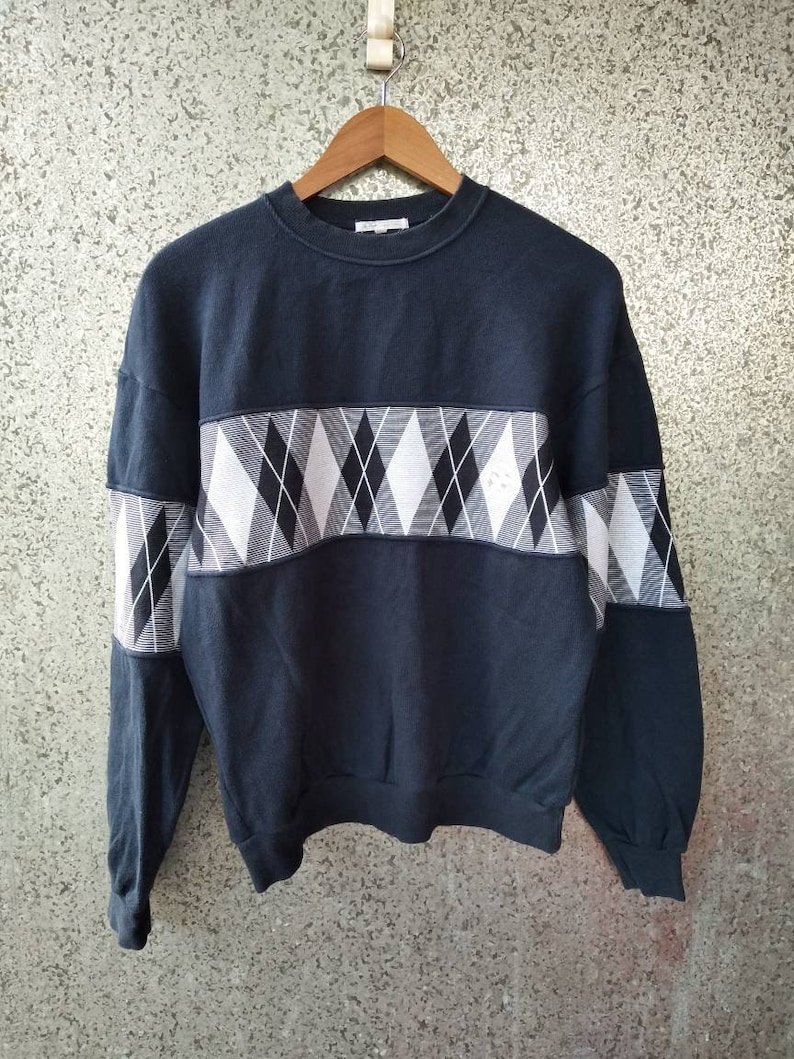 May include: A navy blue sweatshirt with a white and black argyle pattern across the chest and upper sleeves. The sweatshirt is hanging on a wooden hanger against a textured background.