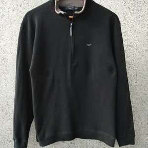 Könnte beinhalten: Schwarzer Langarm-Pullover mit einem halben Reißverschluss und einem kleinen Logo auf der Brust. Der Pullover hängt an einem Holzbügel.