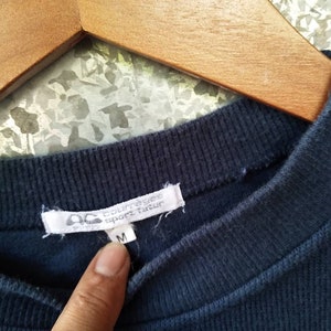 May include: Close-up of a navy blue ribbed crew neck shirt. The white tag displays the brand name "courreges sport futur" and the size "M". The shirt is hanging on a wooden hanger.