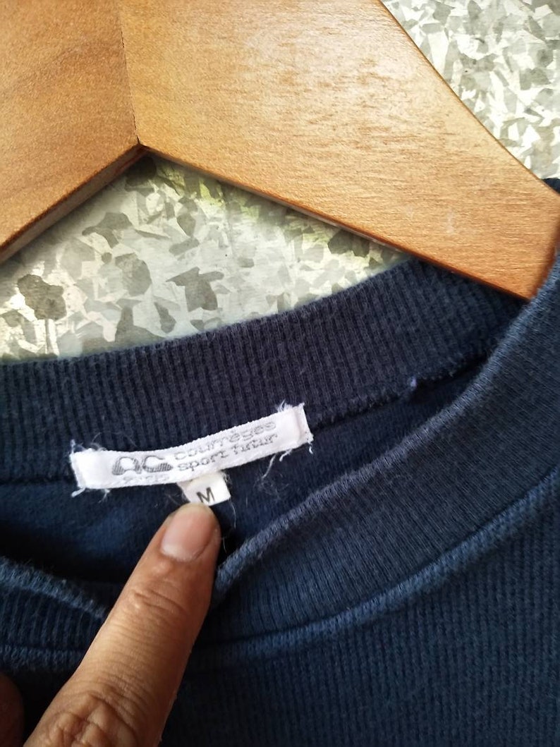 May include: Close-up of a navy blue ribbed crew neck shirt. The white tag displays the brand name "courreges sport futur" and the size "M". The shirt is hanging on a wooden hanger.