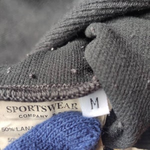May include: Close-up of a dark gray sportswear garment tag. The tag reads "SPORTSWEAR COMPANY" and includes a size "M" label. The fabric appears to be a blend of materials.