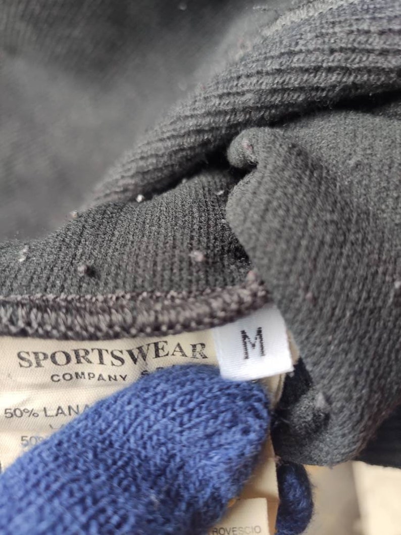 May include: Close-up of a dark gray sportswear garment tag. The tag reads "SPORTSWEAR COMPANY" and includes a size "M" label. The fabric appears to be a blend of materials.