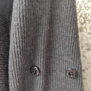 May include: Close-up of a dark gray knit sweater sleeve. The ribbed texture of the fabric is visible, with two black buttons near the cuff. The buttons have a circular design.