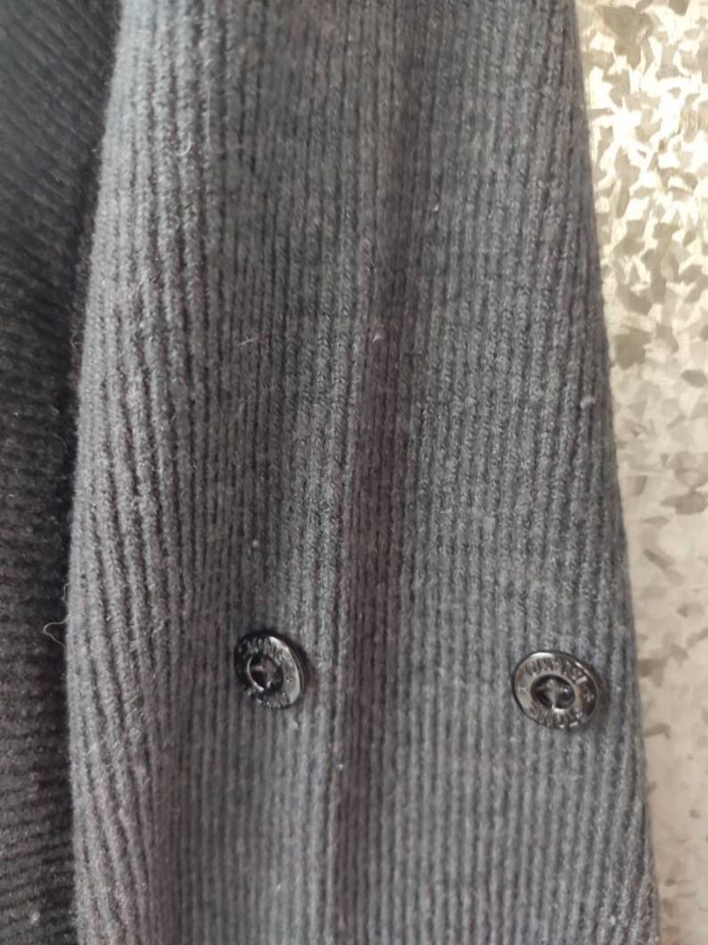 May include: Close-up of a dark gray knit sweater sleeve. The ribbed texture of the fabric is visible, with two black buttons near the cuff. The buttons have a circular design.