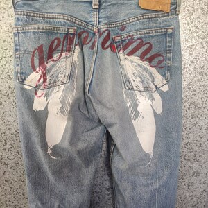 Op de afbeelding: Blauwe denim jeans met witte verenontwerpen en het woord "genuine" in rode letters op de achterzakken.