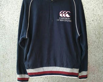 Puede incluir: Jersey de manga larga azul marino con cuello alto y cremallera de cuarto. El jersey presenta ribetes blancos a lo largo de las mangas y un logotipo en el pecho que dice "Canterbury of New Zealand". El dobladillo inferior tiene un diseño a rayas.