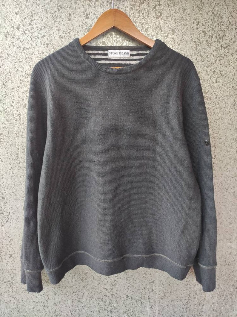 May include: A charcoal gray crew neck sweater with horizontal ribbing. The sweater has long sleeves and a ribbed hem and cuffs. The Stone Island logo is visible on the upper back of the sweater. The sweater is hanging on a wooden hanger.