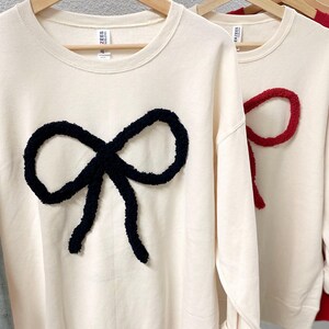 May include: Three cream-colored sweatshirts with a large fuzzy bow on the front. The bows are black, red, and red. The sweatshirts are hanging on wooden hangers.
