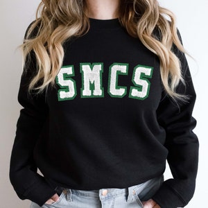 Peut inclure: Sweat-shirt noir avec un graphisme "SMCS" vert et blanc.