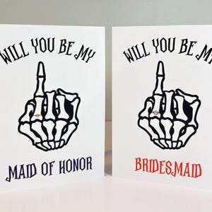 May include: Two white greeting cards featuring a black skeleton hand graphic with a ring on the finger. The text reads WILL YOU BE MY MAID OF HONOR in dark blue and WILL YOU BE MY BRIDESMAID in red, both in a stylised black gothic font.