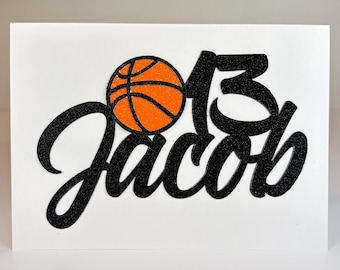 May include: A white card featuring a black glittery script that reads Jacob, with the number 13 in black glitter above the name, and a bright orange glittery basketball icon positioned to the left of the number 13 on the card surface.