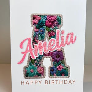 May include: A white greeting card featuring a large letter A filled with intricate, colorful floral embroidery in shades of pink, purple, teal, and green. The name Amelia is written in pink glittery script over the letter, with HAPPY BIRTHDAY below.