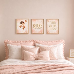 Może przedstawiać: Trzy prostokątne obrazy w złotych ramach przedstawiające ilustrację cheerleaderki, imię Brooke oraz tekst Cheer All Day, have a little sparkle wherever you go, umieszczone nad łóżkiem z różową pościelą i poduszką w kształcie kokardy.