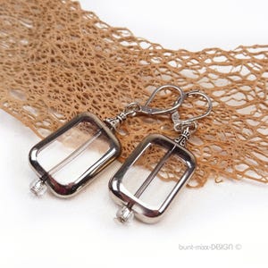 May include: Silver-coloured rectangular earrings with clear glass inserts and a small, clear bead. The earrings have lever-back closures. The background features a textured, brown, net-like material. The earrings are a jewellery item.