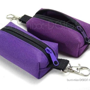 Könnte beinhalten: Zwei violette Reißverschlusstaschen mit schwarzem Besatz und silbernen Schlüsselanhängern. Die Taschen sind aus einem strukturierten Stoff gefertigt und haben ein kleines Etikett mit der Aufschrift "bunt-mix-DESIGN".