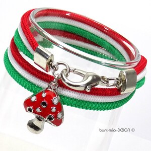 Könnte beinhalten: Ein rotes, weißes und grünes Seil-Armband mit einem silbernen Verschluss und einem roten und weißen Pilz-Anhänger. Das Armband besteht aus drei Seilsträngen, jeder in einer anderen Farbe. Der Pilz-Anhänger ist mit einer silbernen Kette am Verschluss befestigt.