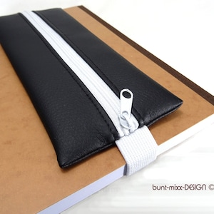 Op de afbeelding: Zwarte leren potlood etui met een witte rits en een wit elastiek. Het etui ligt op een bruin notitieboek.