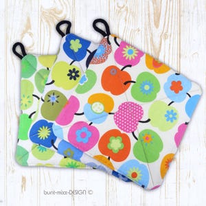 Op de afbeelding: Drie kleurrijke, gewatteerde pannenlappen met zwarte ophanglussen. De pannenlappen hebben een witte achtergrond met een herhaald patroon van gestileerde appels en bloemen in groen, roze, oranje en blauw. De tekst "bunt-mixx-DESIGN" is zichtbaar.