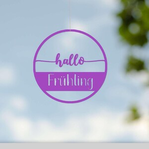 Könnte beinhalten: Eine lila kreisförmige Dekoration mit den Worten "hallo Frühling" in einer Schreibschrift. Die Dekoration ist an einem dünnen Faden vor einem verschwommenen Hintergrund aus Himmel und Laub aufgehängt.