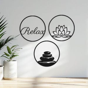May include: Three black metal wall art pieces. One circle features the word "Relax" in cursive. Another circle displays a lotus flower design. The third circle shows stacked stones. All are mounted on a white wall.