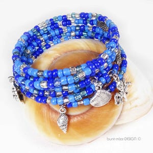 Può includere: Bracciale a più fili con perline blu e argento, ornato da ciondoli a forma di conchiglia. Il bracciale è avvolto attorno a una conchiglia, creando un tema costiero. Le perline sono piccole e ben distanziate.