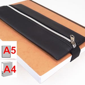 Può includere: Astuccio per matite nero con cerniera, con una cerniera argentata e un cinturino nero. L'astuccio è appoggiato su un quaderno marrone con il testo "A5" e "A4" stampato sulle pagine.