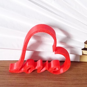 Op de afbeelding: Een rode hartvormige koekjessnijder met het woord "love" in cursief geschreven. De snijder is gemaakt van een flexibel materiaal en ligt op een houten oppervlak. De achtergrond bevat wit papier en een hint van een boek.