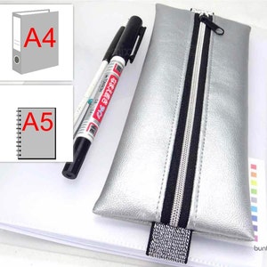 Op de afbeelding: Een zilveren etui met rits, met een zwarte rits en een glimmende zilveren rits-trekker. Het etui ligt op een wit oppervlak naast een zwart-witte markeerstift.