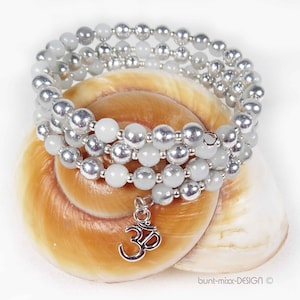 Könnte beinhalten: Ein mehrreihiges Armband mit silbernen Perlen und hellen Perlen, möglicherweise Jade. Das Armband liegt auf einer Muschelschale und hat einen silbernen Anhänger mit dem Om-Symbol.