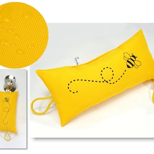 Könnte beinhalten: Gelber Türstopper in Form eines Kissens mit einem schwarzen Bienendesign und einer schwarzen gepunkteten Linie, die hinter ihm herläuft. Der Türstopper hat eine gelbe elastische Schlaufe.