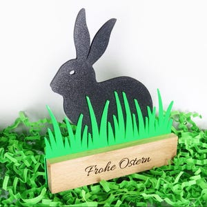 Op de afbeelding: Zwart silhouet van een paashaas met een textuur, tegen felgroen gras. Het konijn is gemonteerd op een houten voet met de woorden "Frohe Ostern" gegraveerd. De achtergrond is wit.