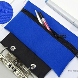 Könnte beinhalten: Ein blaues Federmäppchen mit schwarzem Reißverschluss und einem kleinen schwarzen Etikett mit der Aufschrift "bunt". Darunter befindet sich ein schwarzes Federmäppchen. Ein weißer Stift und ein grauer Marker liegen auf dem blauen Federmäppchen. Das Federmäppchen ist aus wasserdichtem Material.
