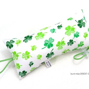 Könnte beinhalten: Ein weißer Stoff-Türstopper mit grünem Kordelverschluss. Der Stoff ist mit einem Muster aus grünen vierblättrigen Kleeblättern bedruckt. Der Türstopper hat ein Etikett mit der Aufschrift "Zeige bunt!"