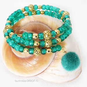 May include: A wrap bracelet with three strands of teal beads and gold-coloured accents. The bracelet features decorative gold beads and a teal pom-pom charm. The bracelet is displayed on a seashell.