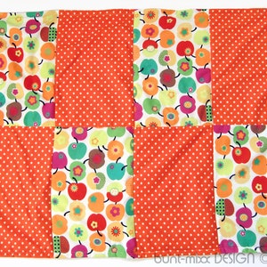 May include: A patchwork quilt with a square design. The quilt is made of four different fabrics. Two of the fabrics have a white background with a pattern of green and red apples. The other two fabrics are orange with white polka dots.