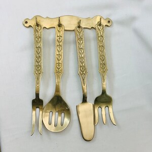 May include: A set of four gold-toned metal utensils with ornate designs hanging on a wall-mounted rack. The utensils include a fork, a spoon, a knife, and a serving spoon.
