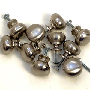 Peut inclure: Un ensemble de dix boutons de cabinet argentés de forme ronde légèrement bombée. Les boutons ont une finition brossée et sont fixés à des vis.