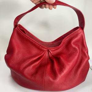 Peut inclure: Un sac hobo en cuir rouge avec une seule bandoulière. Le sac présente un design froncé à la base et une surface texturée. Le sac est tenu par une main.