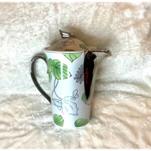 May include: White ceramic teapot with a silver lid and black handle. The teapot features a floral design with green leaves and white flowers.