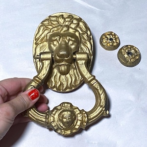 May include: A gold-coloured lion head door knocker with a ring handle. The knocker features a detailed lion's face with a mane and a ring handle below. Two small, round, gold-coloured mounting plates are also visible.