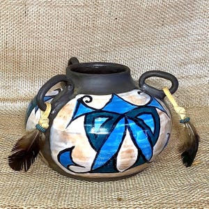 May include: A brown ceramic vase with two handles and a blue, green and white floral design. The vase has two leather straps with feathers attached.