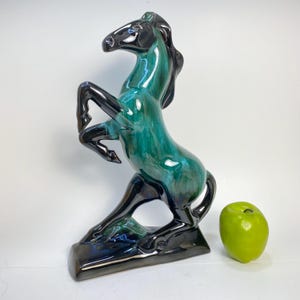 May include: A ceramic horse statue in a rearing pose, with a glossy teal and black glaze. The sculpture is on a rectangular base. A green apple is to the right of the horse.