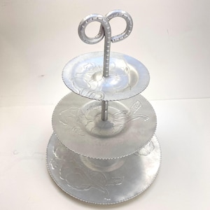 May include: A three-tiered aluminium serving stand with a floral design. The stand has a decorative handle and is ideal for serving snacks or desserts.