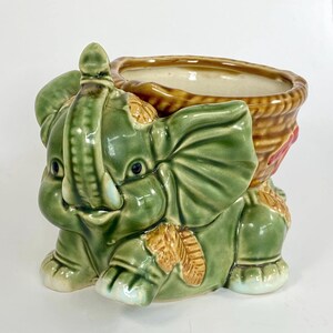Peut inclure: Un pot de fleurs en céramique vert en forme d'éléphant avec un panier en osier brun sur le dos. L'éléphant a un visage souriant et de grandes oreilles. Le pot est parfait pour les petites plantes ou les succulentes.