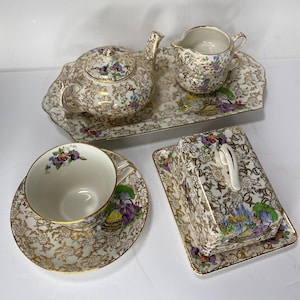May include: A vintage tea set comprising a teapot, creamer jug, cup and saucer, butter dish, and serving tray. The set has a gold floral pattern with purple, yellow, and green accents. Gold trim edges.