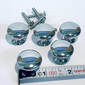 May include: Five chrome cabinet knobs with a round top and a smaller round base. The knobs are arranged in a semi-circle with a measuring tape in the foreground. The tape reads 9 ft.